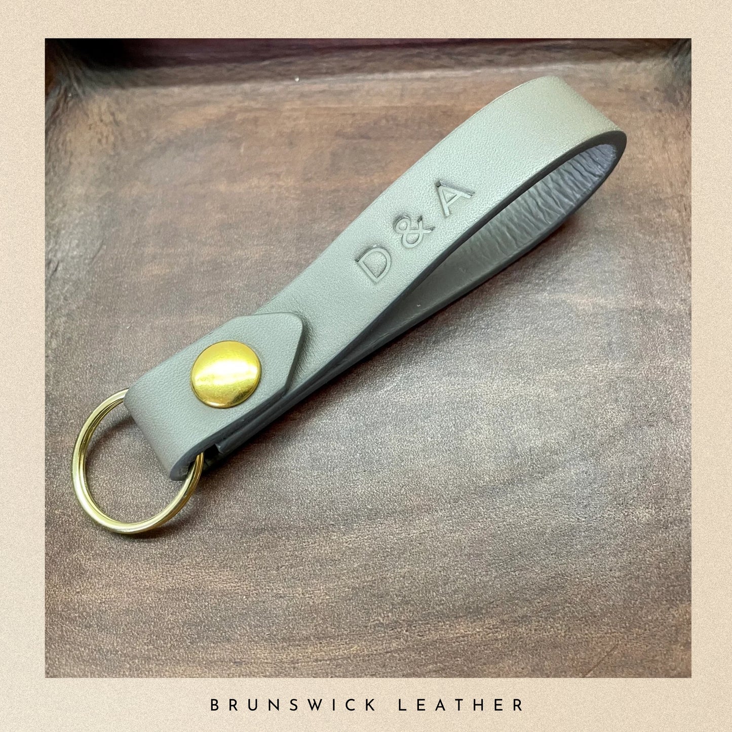 Grey leather key fob with gold accent on a wooden surface, branded 'Brunswick Leather'.