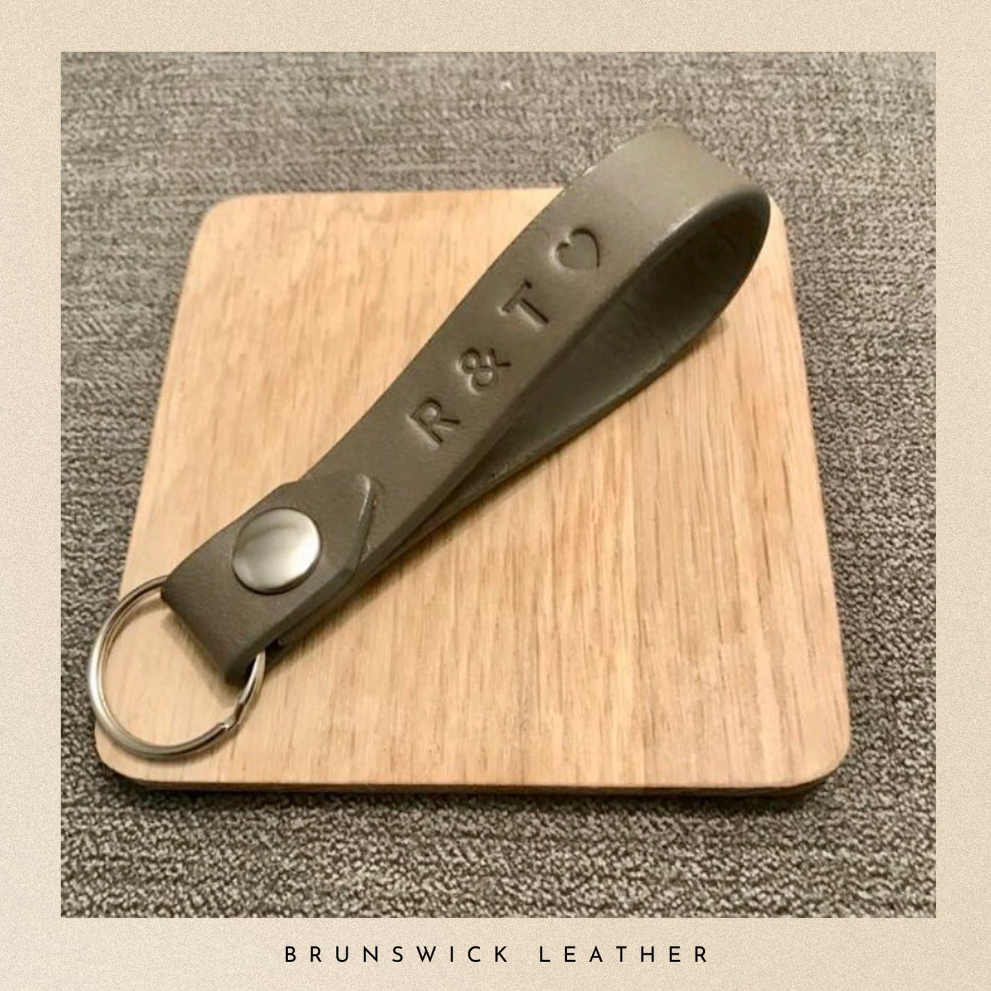 Grey leather keychain with 'R & T' engraving on a wooden board, branded 'Brunswick Leather'.