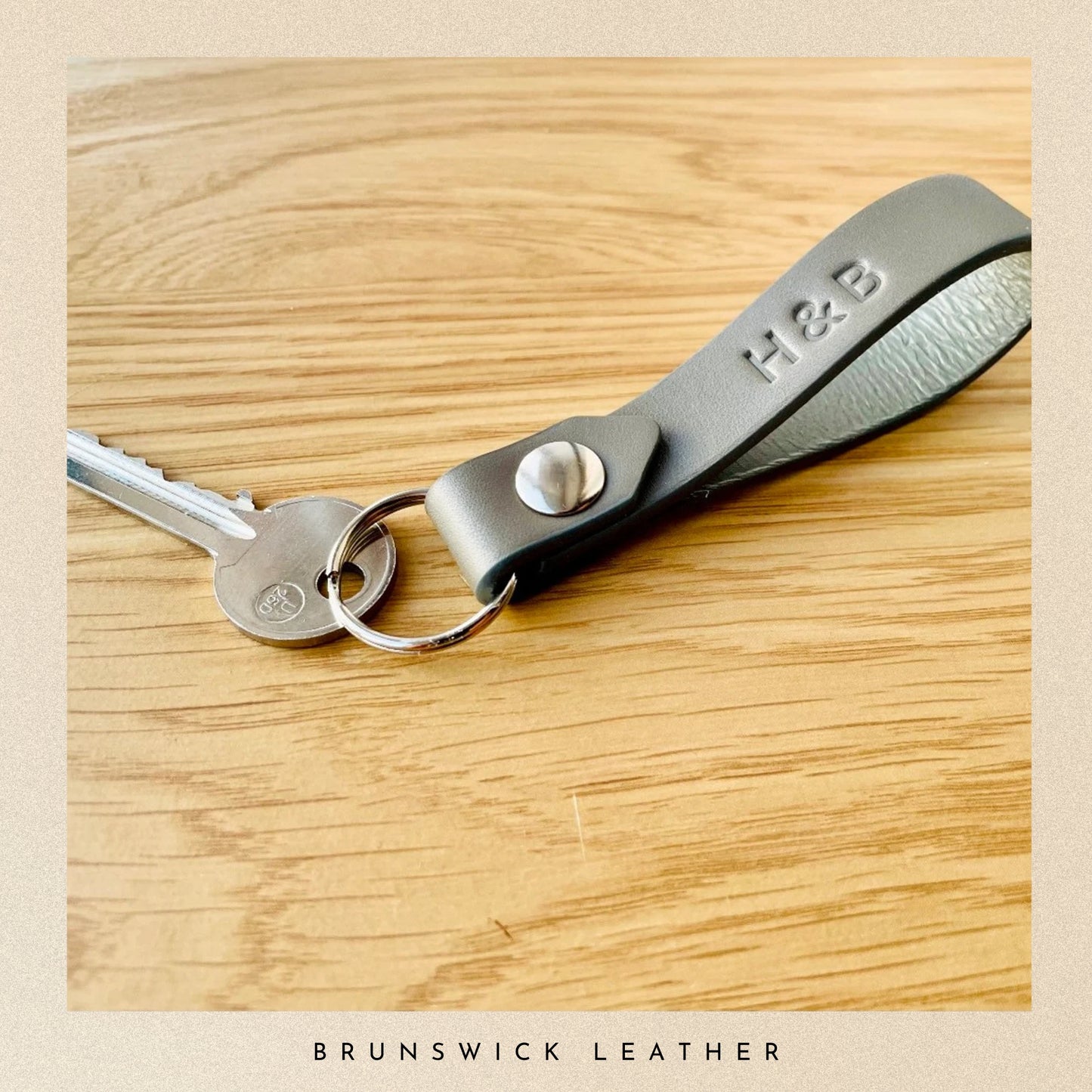 Keychain with grey leather strap and metal clasp on a wooden surface, branded 'H&B'.