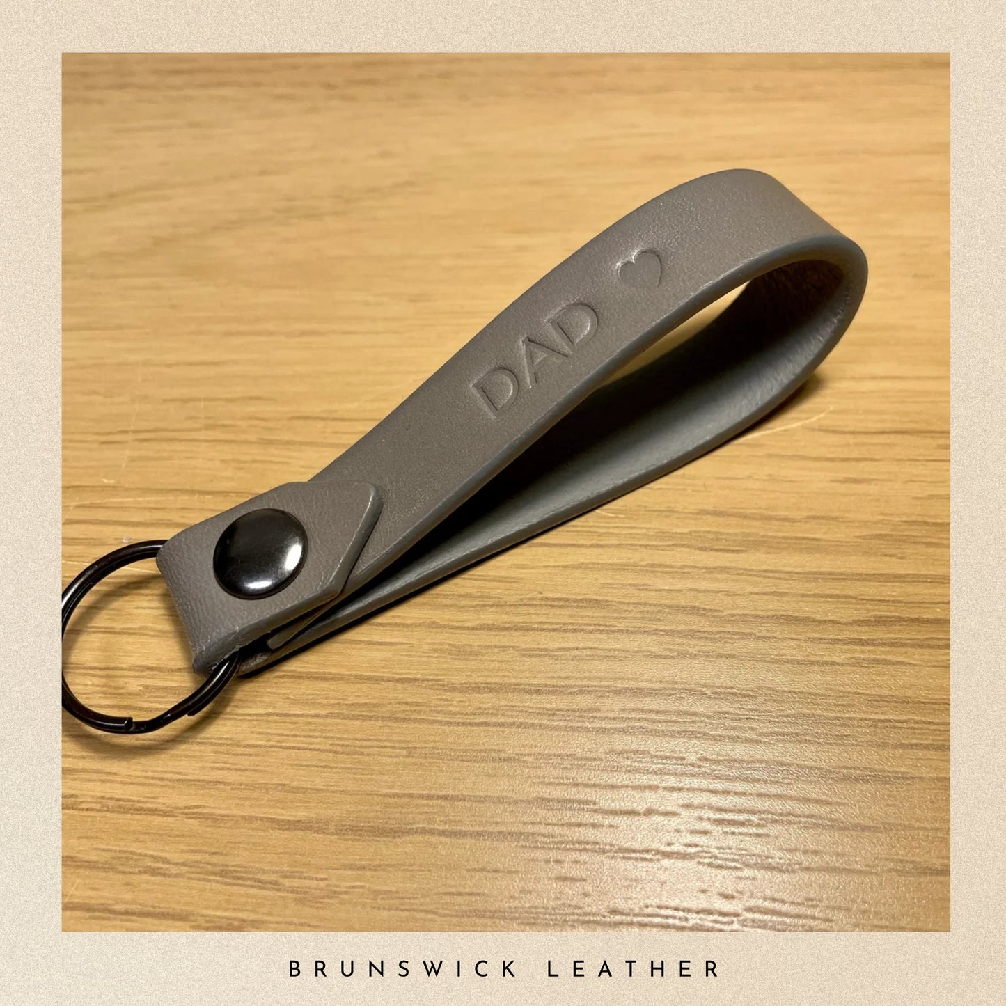Grey leather keychain with 'DAD' engraving on a wooden surface, branded 'Brunswick Leather'.