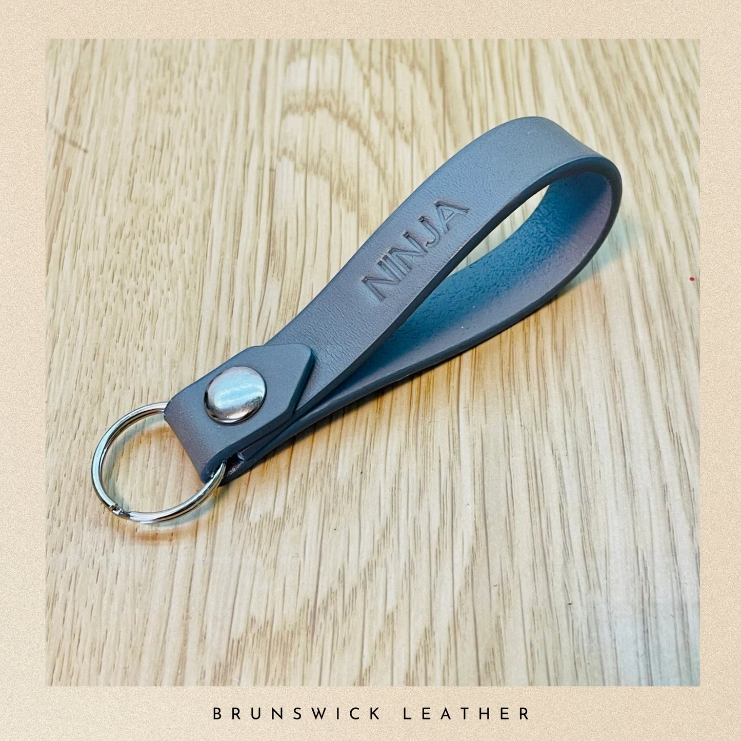 Grey leather keychain with 'NINJA' engraving on a wooden surface, branded 'Brunswick Leather'.
