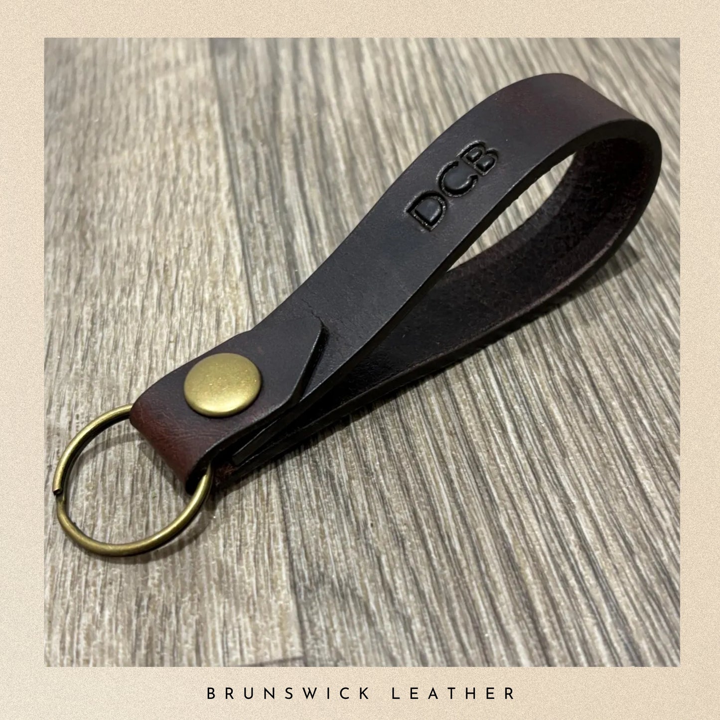 Brown leather keychain with brass ring and 'DCB' engraving on a wooden surface, branded 'Brunswick Leather'.