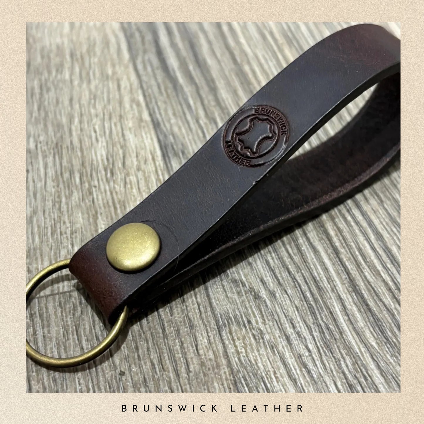 Brown leather keychain with gold accent on a wooden surface, branded 'Brunswick Leather'.