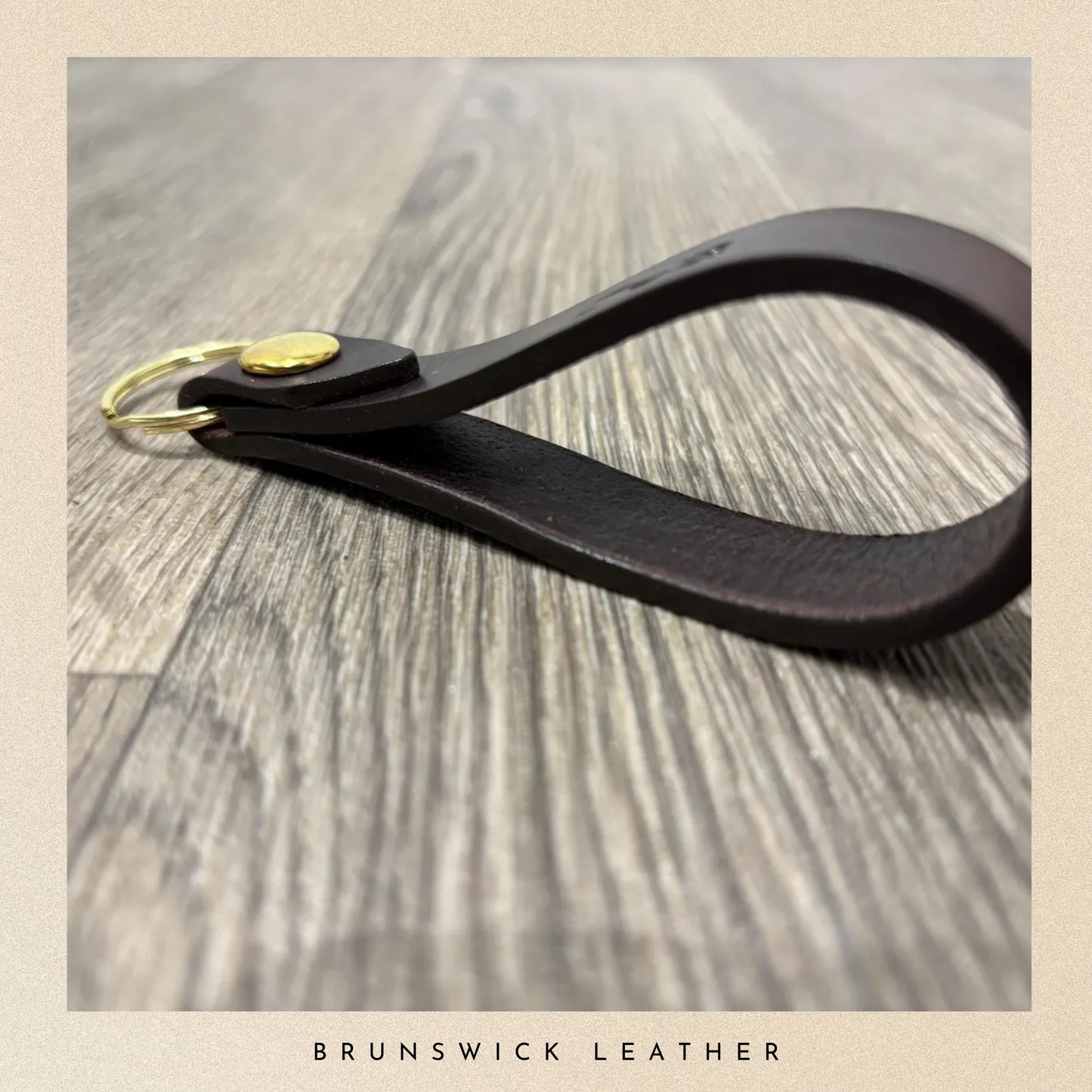 Black leather keychain with gold accent on a wooden surface, branded 'Brunswick Leather'.