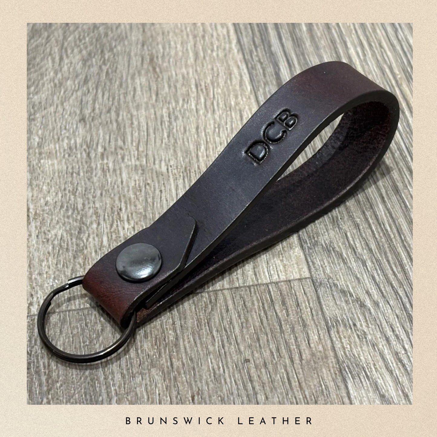 Brown leather keychain with 'DCB' branding on a wooden surface, branded 'Brunswick Leather'.