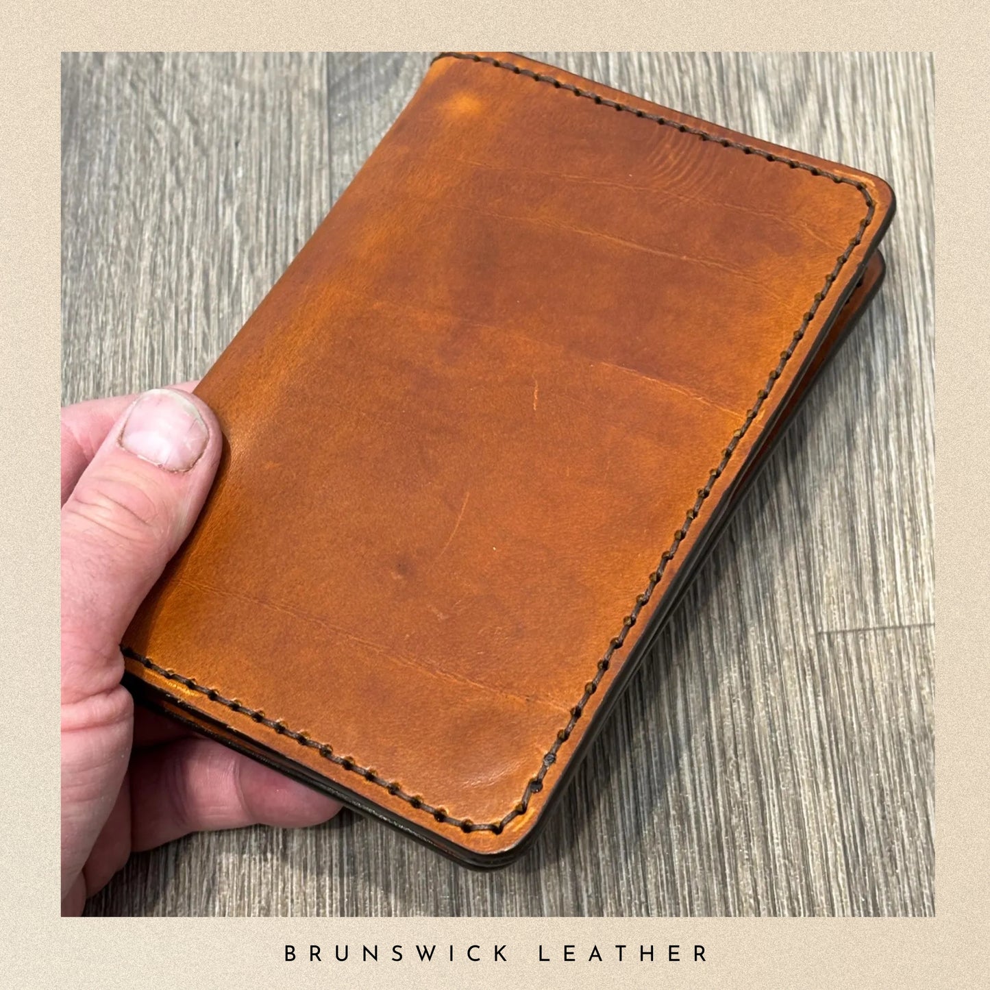 Brown leather wallet held by a hand on a wooden surface with 'Brunswick Leather' branding.