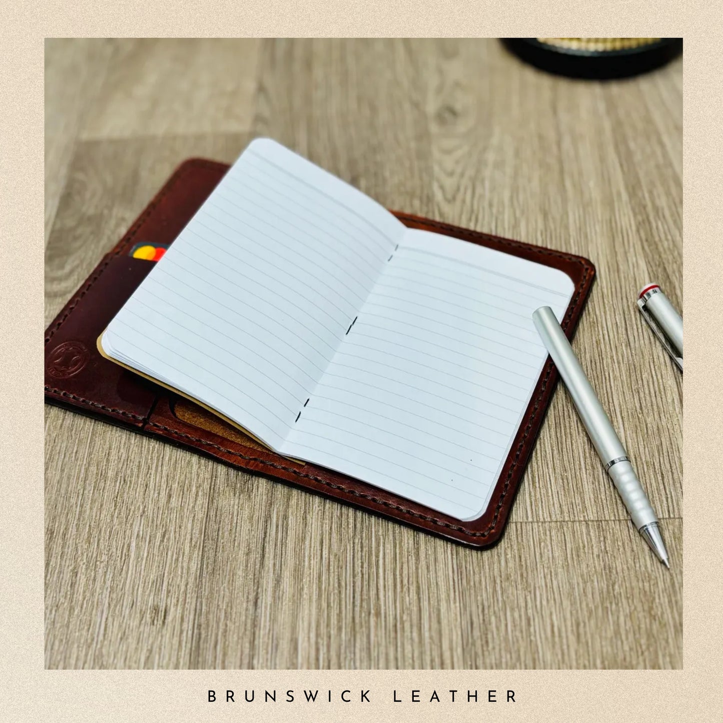 Open notebook with lined pages inside, placed on a wooden surface with a pen beside it, branded 'Brunswick Leather'.