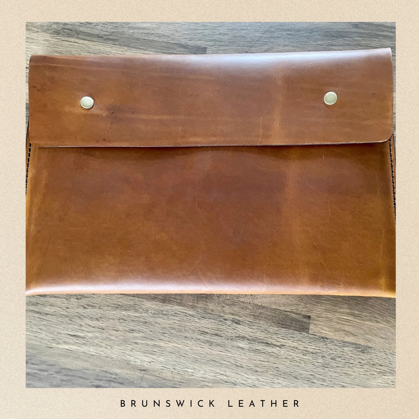Brown leather clutch with silver studs on a wooden surface, branded 'Brunswick Leather'.