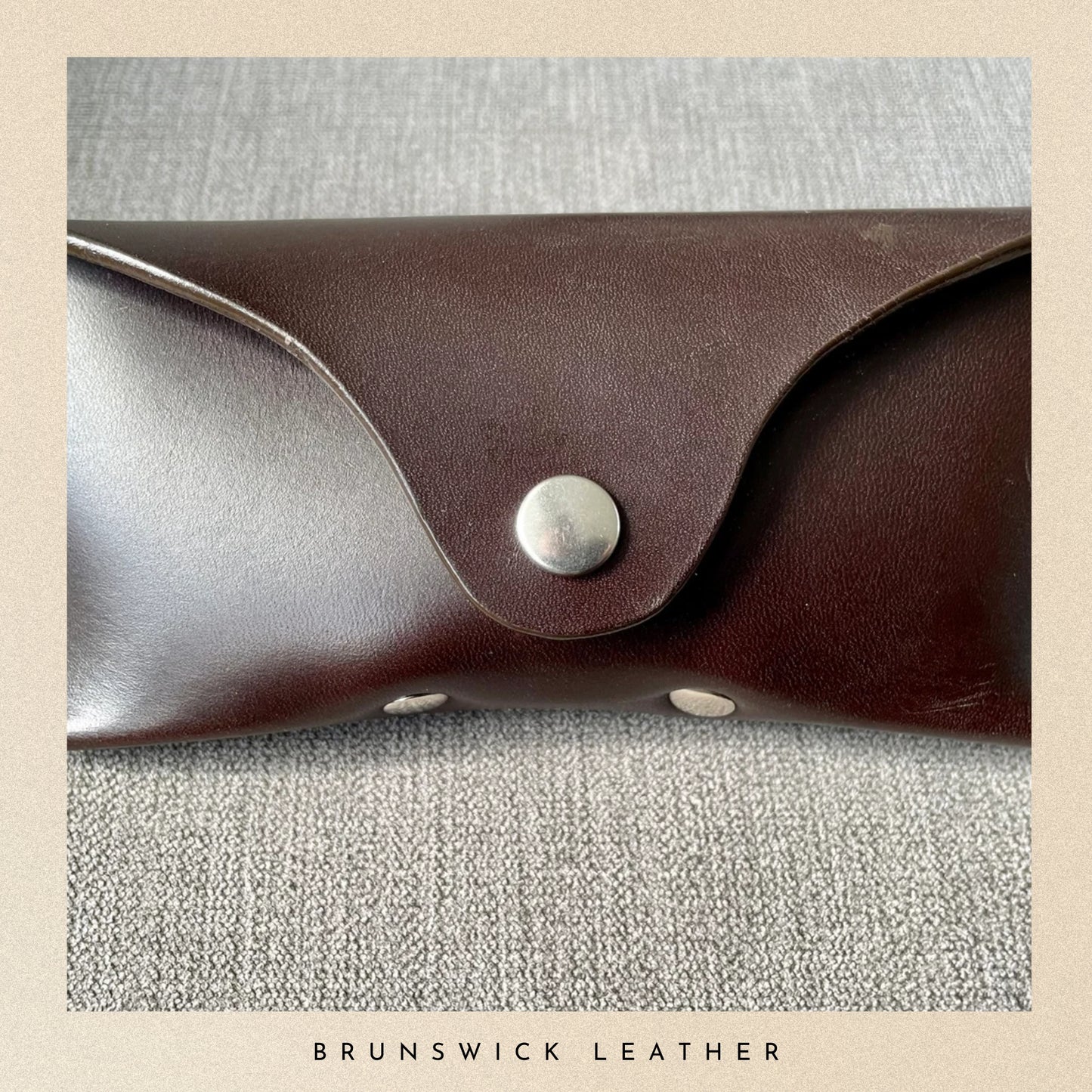 Brown leather case with a silver button on a textured gray background, branded 'Brunswick Leather'.