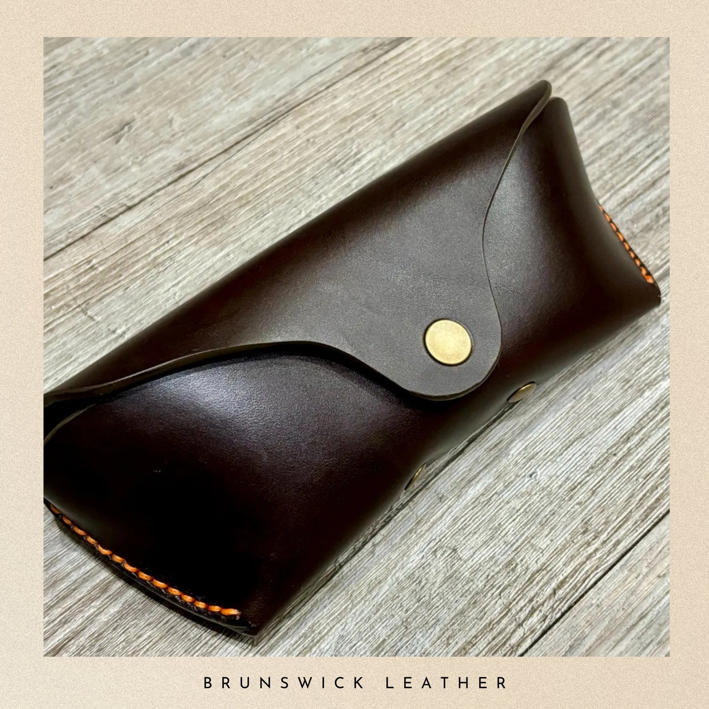 Brown leather case with gold button on a wooden surface, branded 'Brunswick Leather'.
