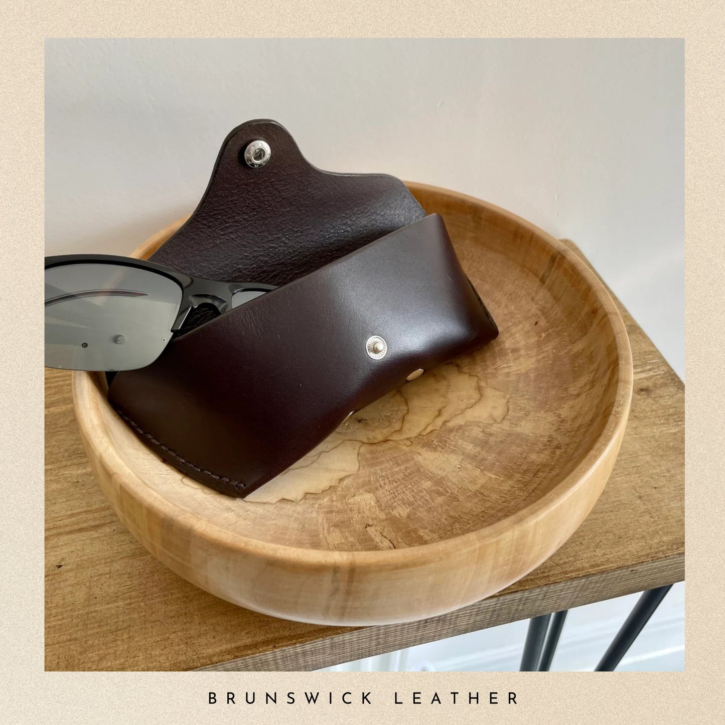 Brown leather sunglasses case on a wooden bowl with 'Brunswick Leather' branding.