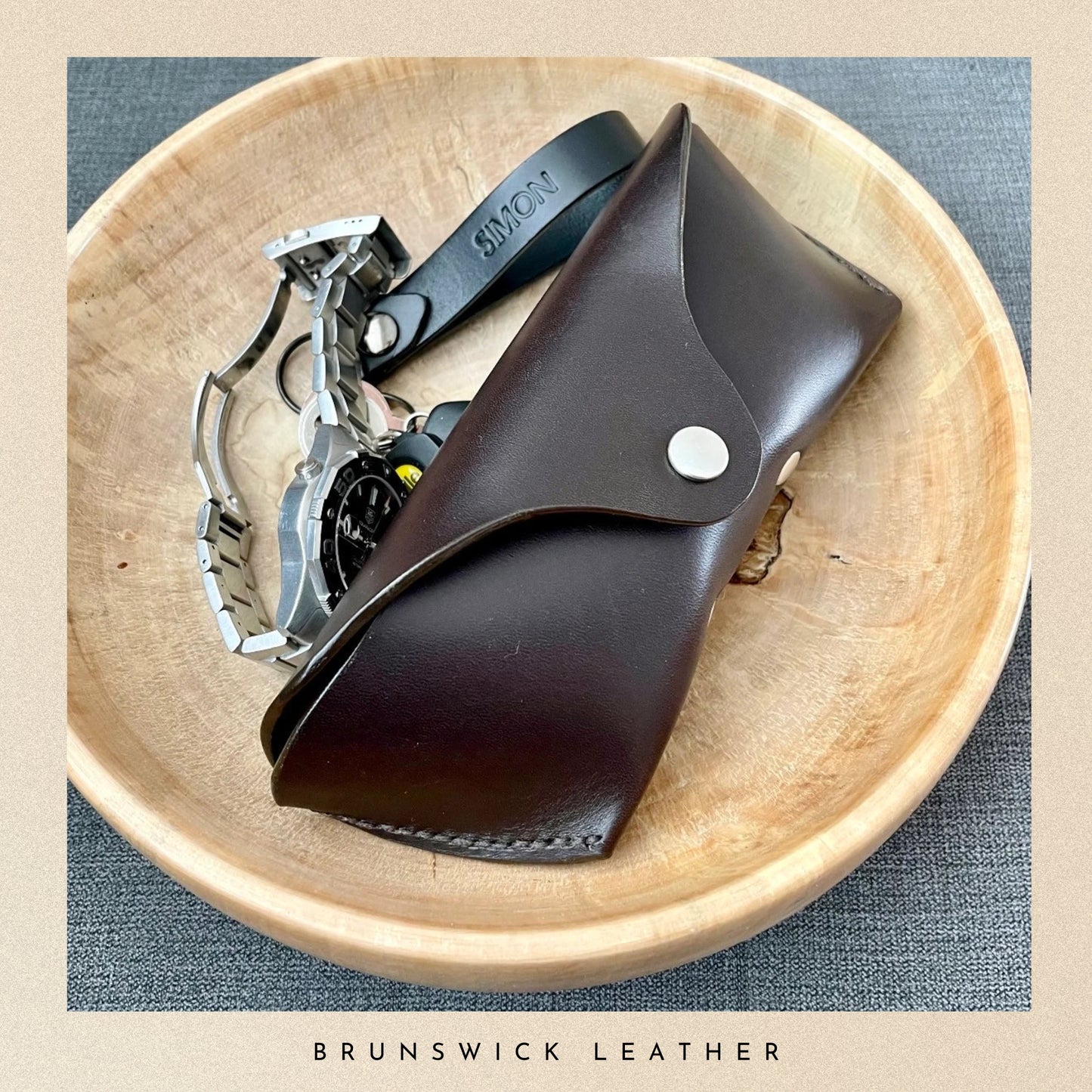 Brown leather pocket knife and black belt on a wooden surface with 'Brunswick Leather' branding.