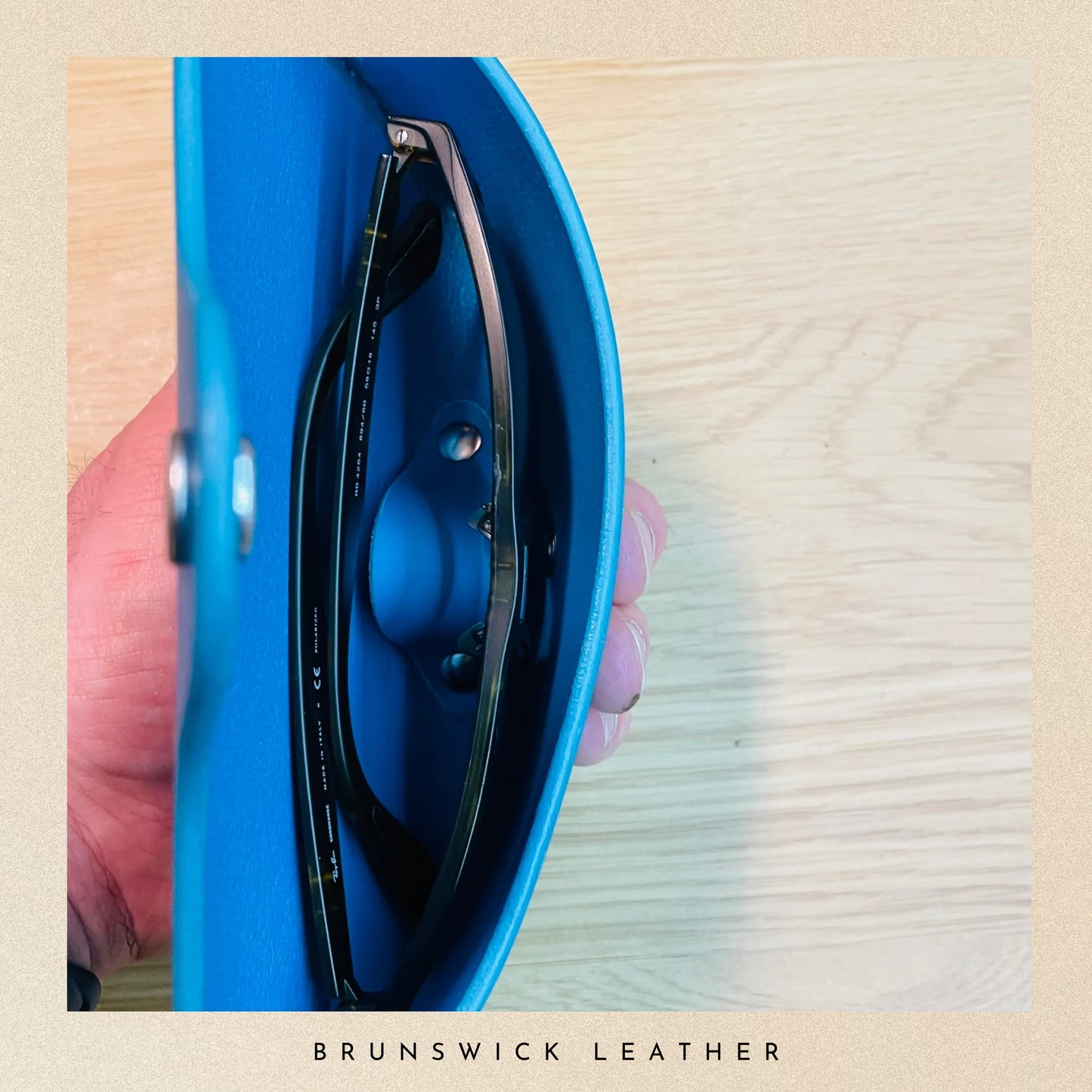 Blue leather case with sunglasses held by a hand on a wooden surface, branded 'Brunswick Leather'.