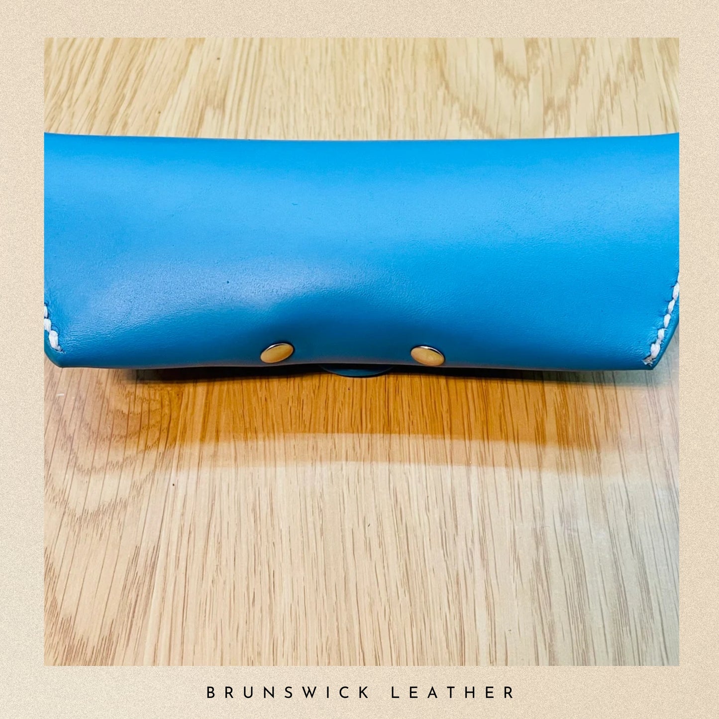 Blue leather wallet with gold buttons on a wooden surface, branded 'Brunswick Leather'.