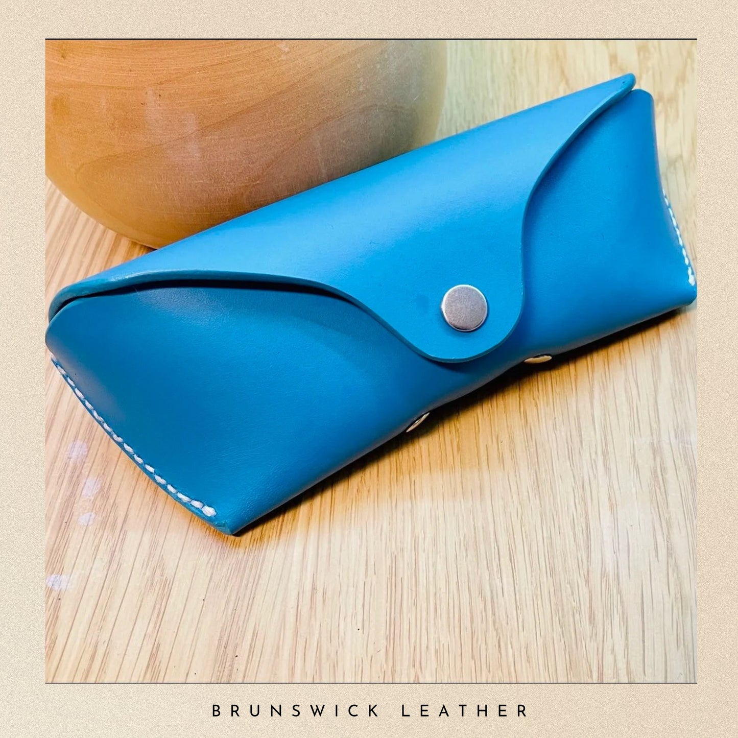 Blue leather case with a metallic button on a wooden surface, branded 'Brunswick Leather'.
