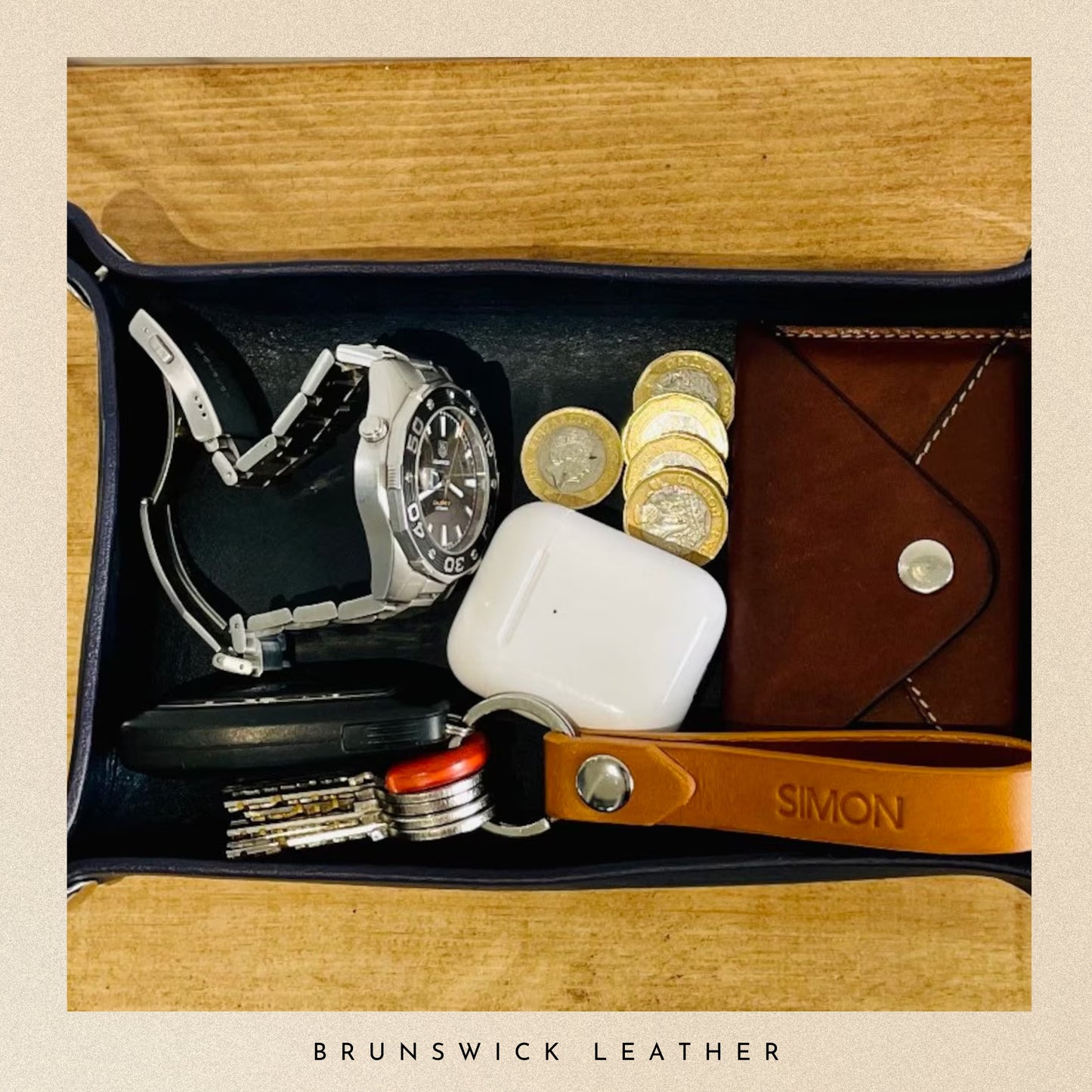 Open leather wallet with coins, watch, keys, and airpods on a wooden surface, branded 'Brunswick Leather'.