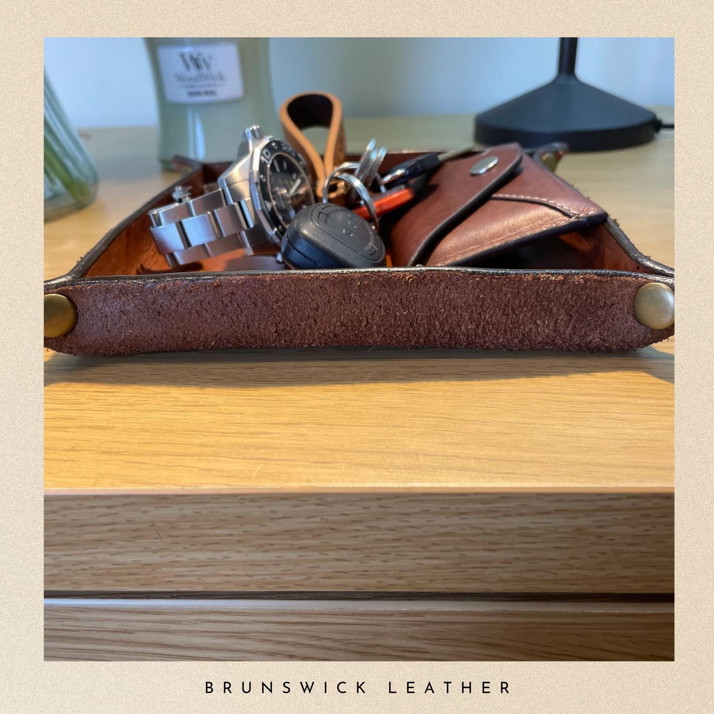 Brown leather key holder with keys and a watch on a wooden surface, branded 'Brunswick Leather'.