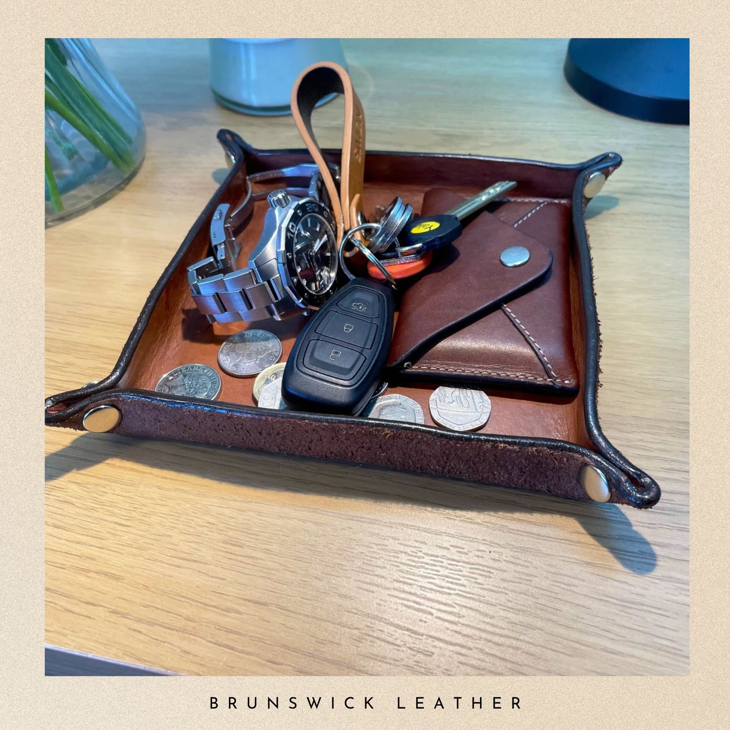 Brown leather tray with keys, wallet, and coins on a wooden surface, branded 'Brunswick Leather'.