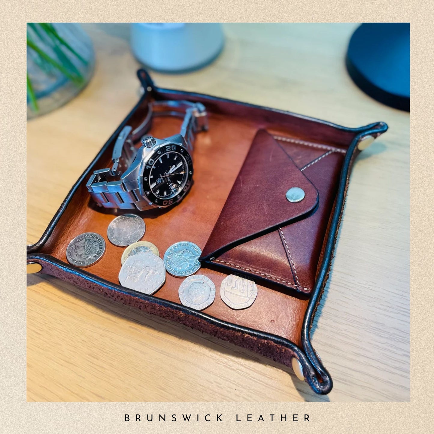 Brown leather tray with coins, watch, and wallet on a wooden surface, branded 'Brunswick Leather'.