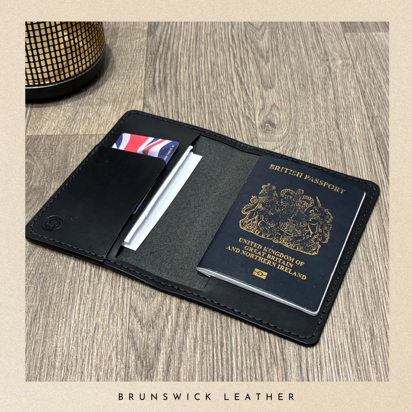 Black leather passport holder with a British passport on a wooden surface, branded 'Brunswick Leather'.