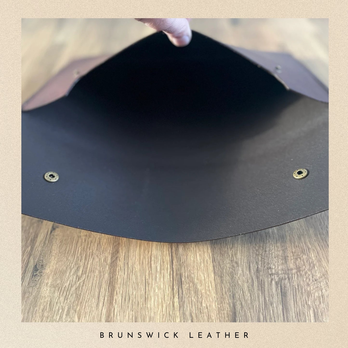 Black leather flap with gold studs on a wooden surface, branded 'Brunswick Leather'.
