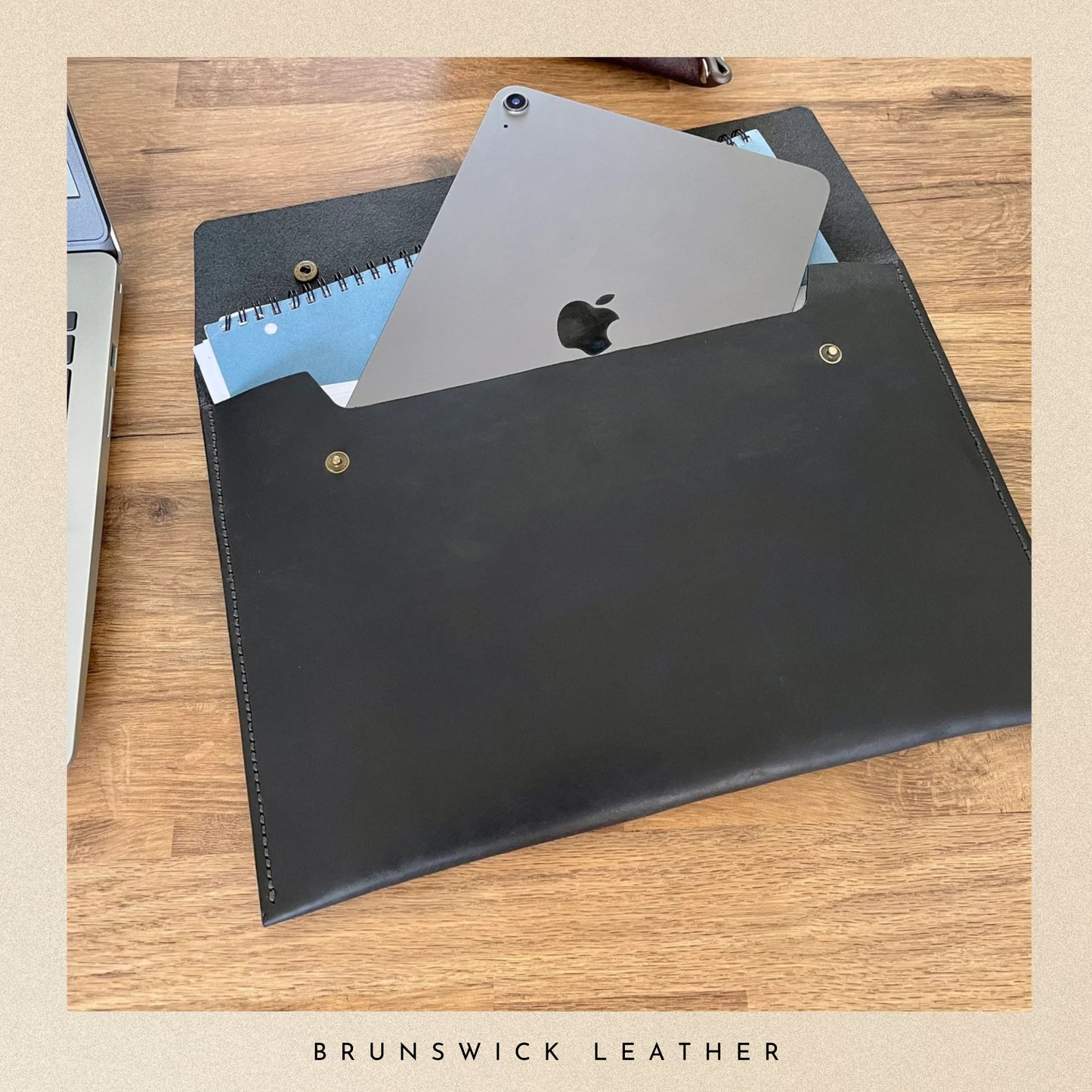 Black leather sleeve with Apple product on a wooden surface, branded 'Brunswick Leather'.