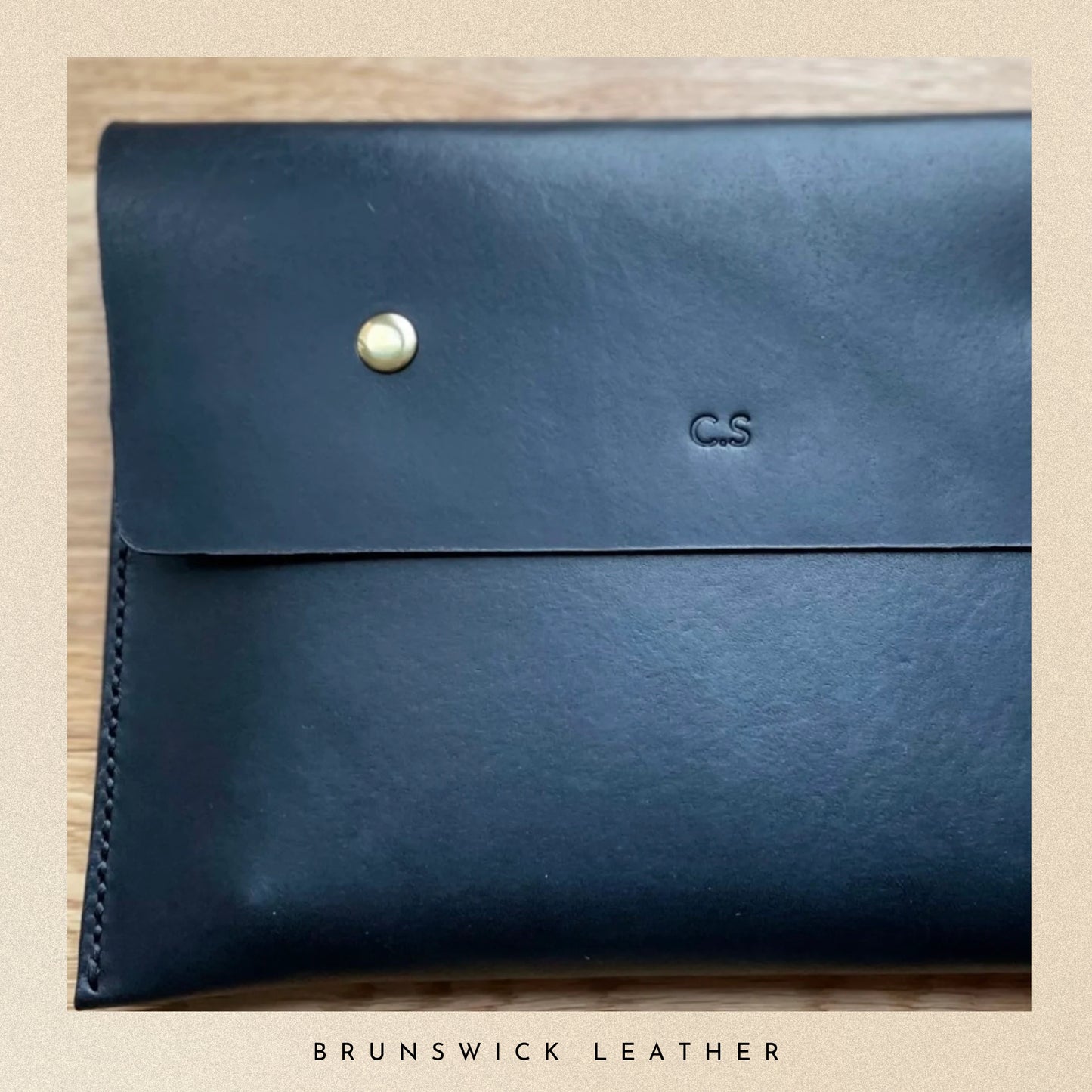 Blue leather clutch with a gold button and initials on a wooden surface, branded 'Brunswick Leather'.