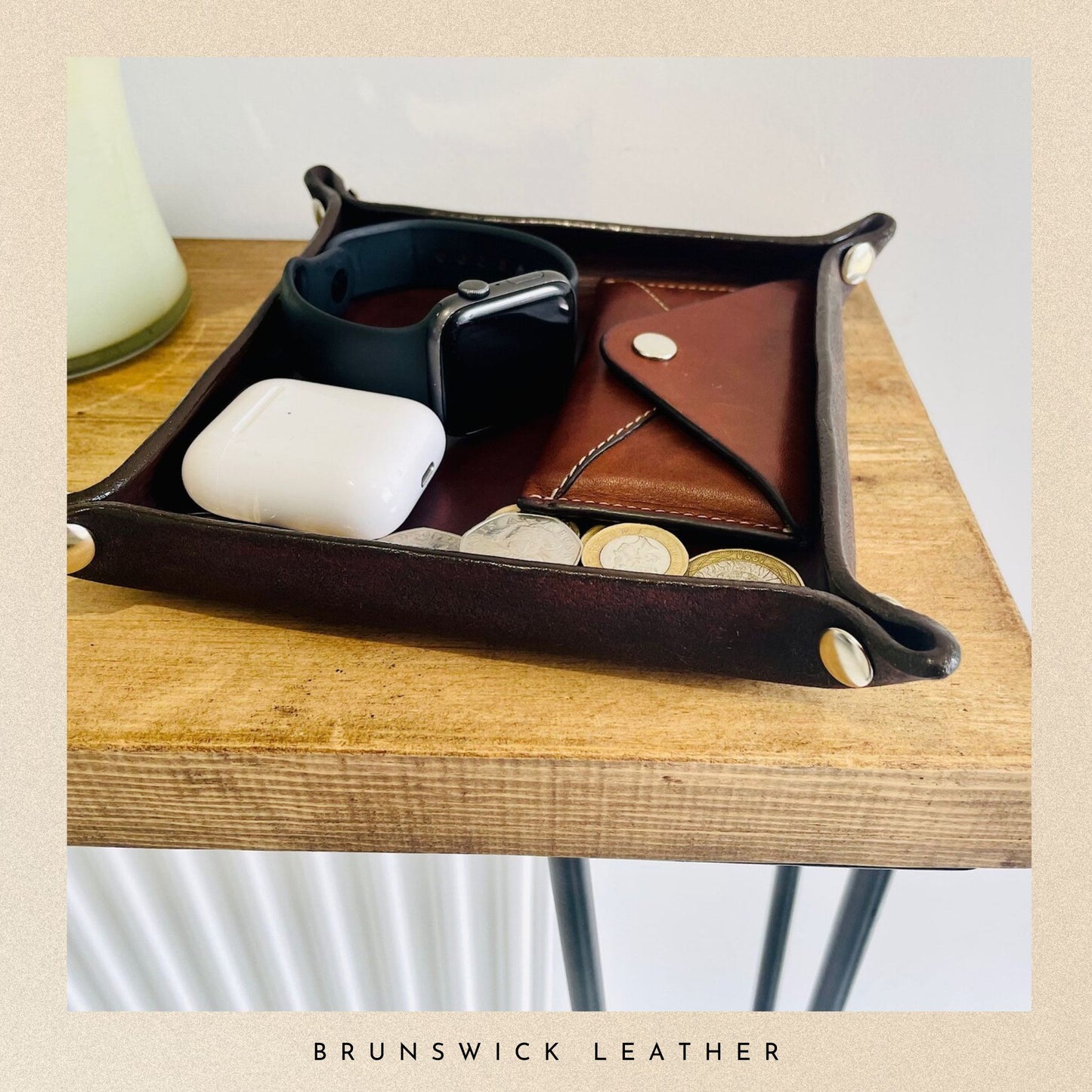 Brown leather tray with sunglasses, coins, and a phone on a wooden surface, branded 'Brunswick Leather'.