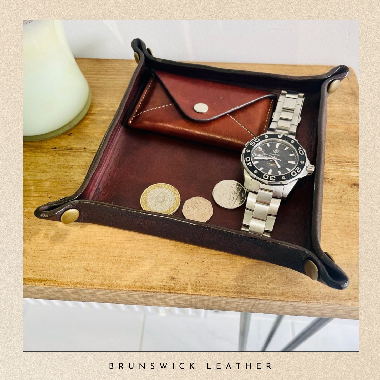 Leather tray with a watch, coins, and wallet on a wooden surface, branded 'Brunswick Leather'.