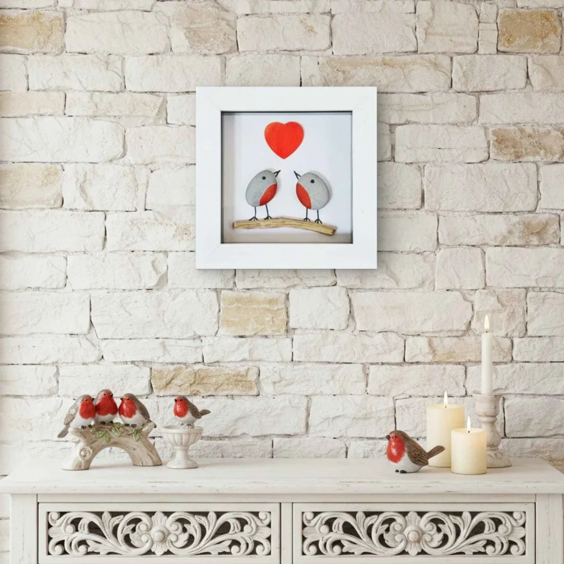 A square white-framed wall art piece hangs on a light stone wall above a white carved console table. The artwork shows two small robins facing each other beneath a bright red heart, with a wooden branch behind them.