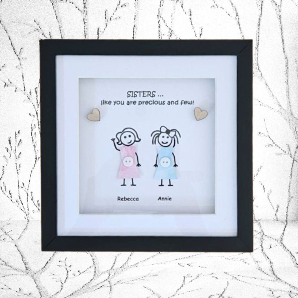 A personalised black shadow box frame with a white mat, featuring a hand-drawn illustration of two sisters in pink and blue dresses with the text 'SISTERS .