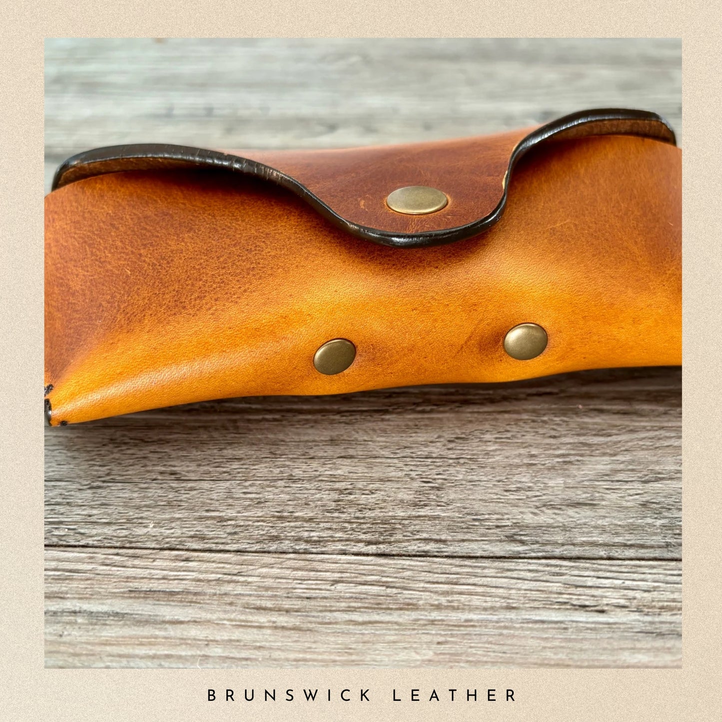 Brown leather case with gold studs on a wooden surface, branded 'Brunswick Leather'.