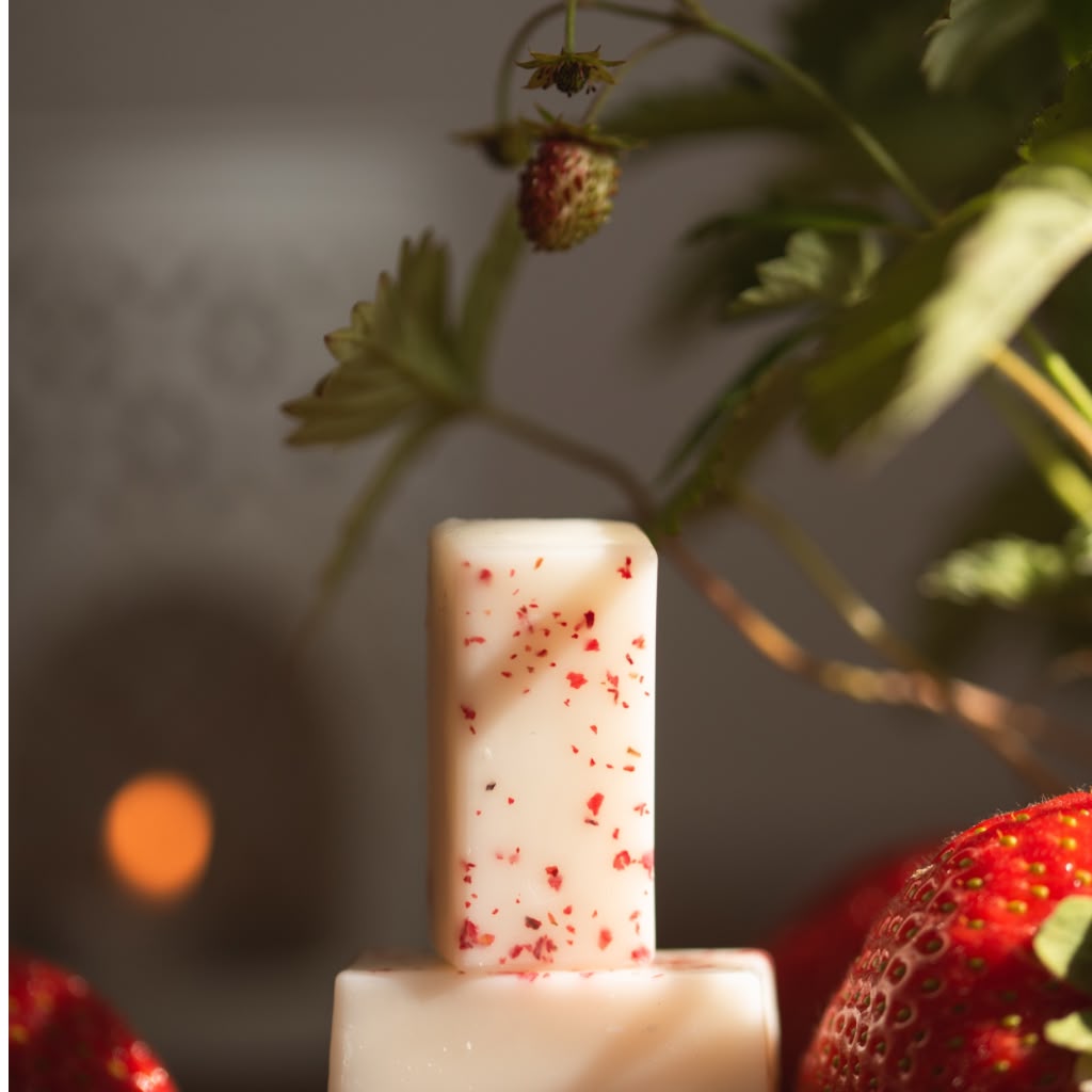 A rectangular wax melts with red strawberry speckles, displayed on a matching base with fresh strawberries and green leaves in a soft-focus background.