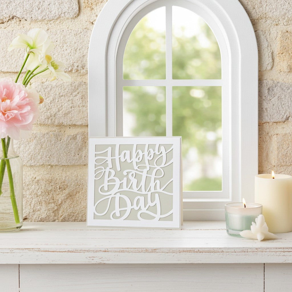 A square white 'Happy Birthday' greeting card displayed on a white wooden mantel in a styled home setting. The scene includes a brick wall background, a white arched window, a vase of pink flowers, and two lit candles.