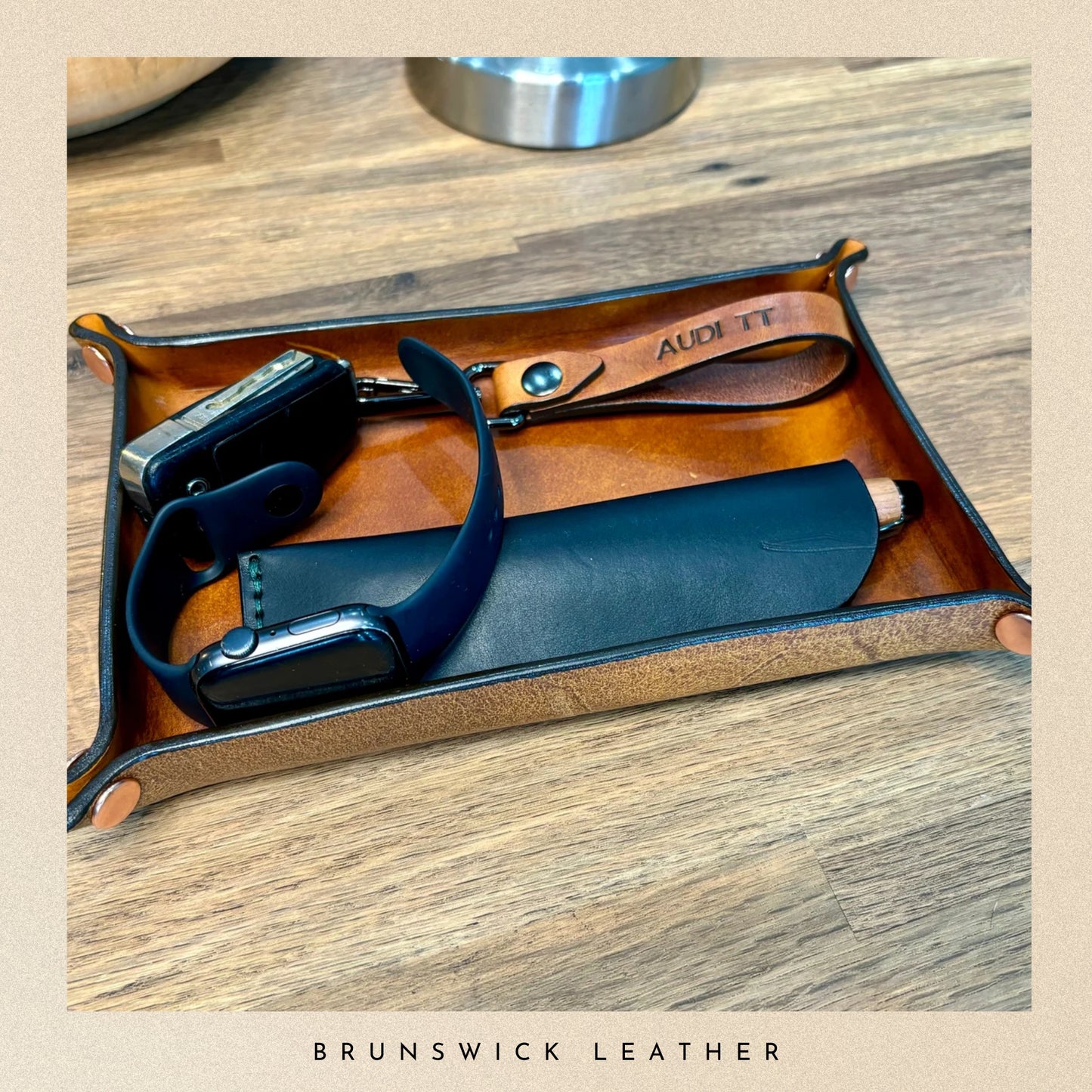 Leather tray with sunglasses and other items on a wooden surface, branded 'Brunswick Leather'.