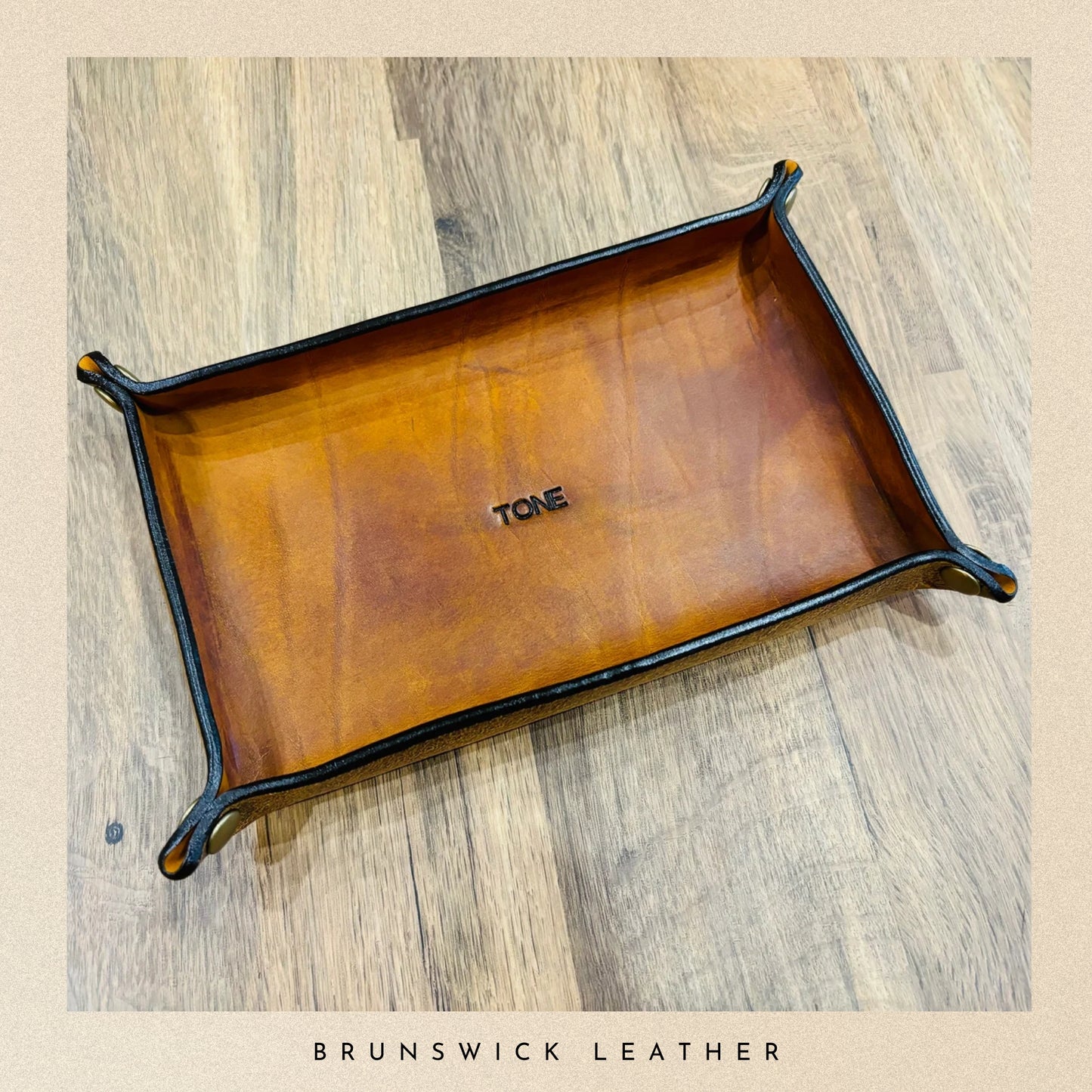 Brown leather tray with 'TONE' engraving on a wooden surface, branded 'Brunswick Leather'.