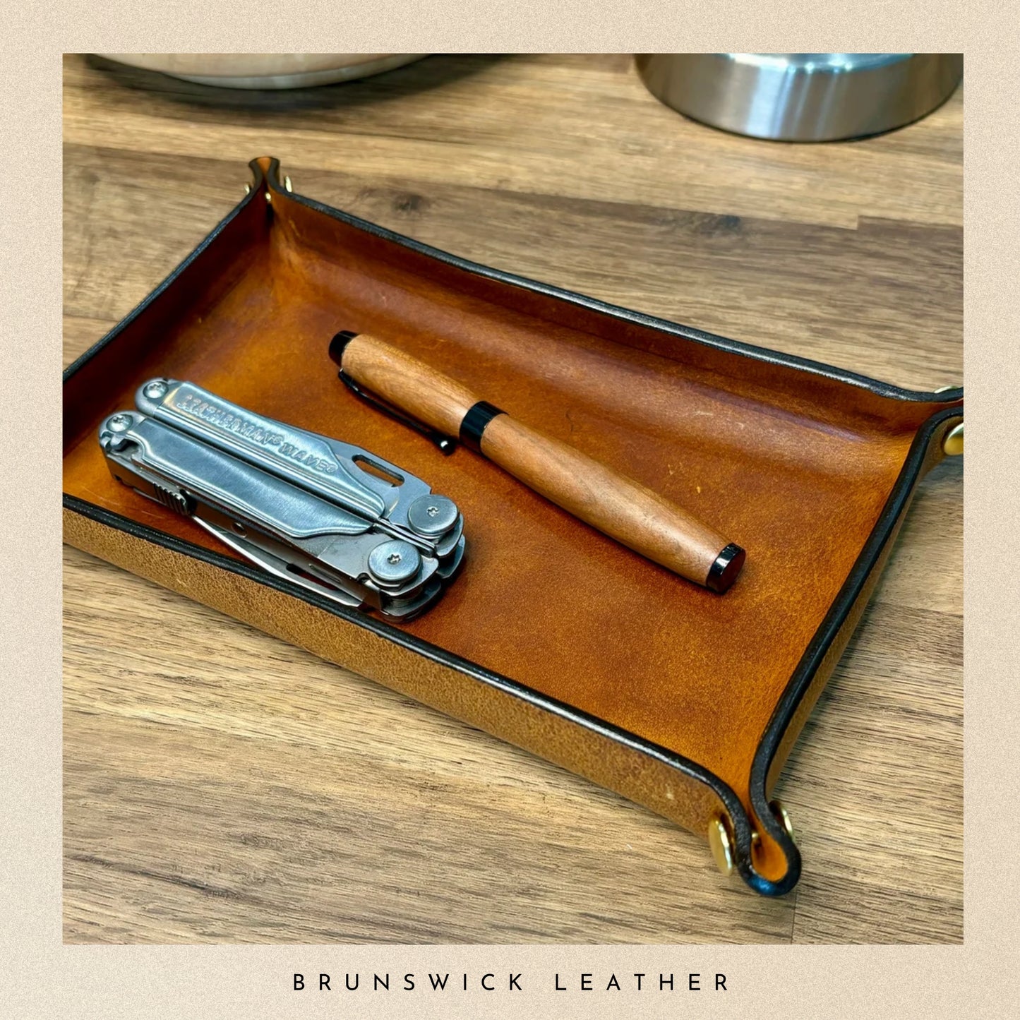 Brown leather tray with a multitool and pen on a wooden surface, branded 'Brunswick Leather'.