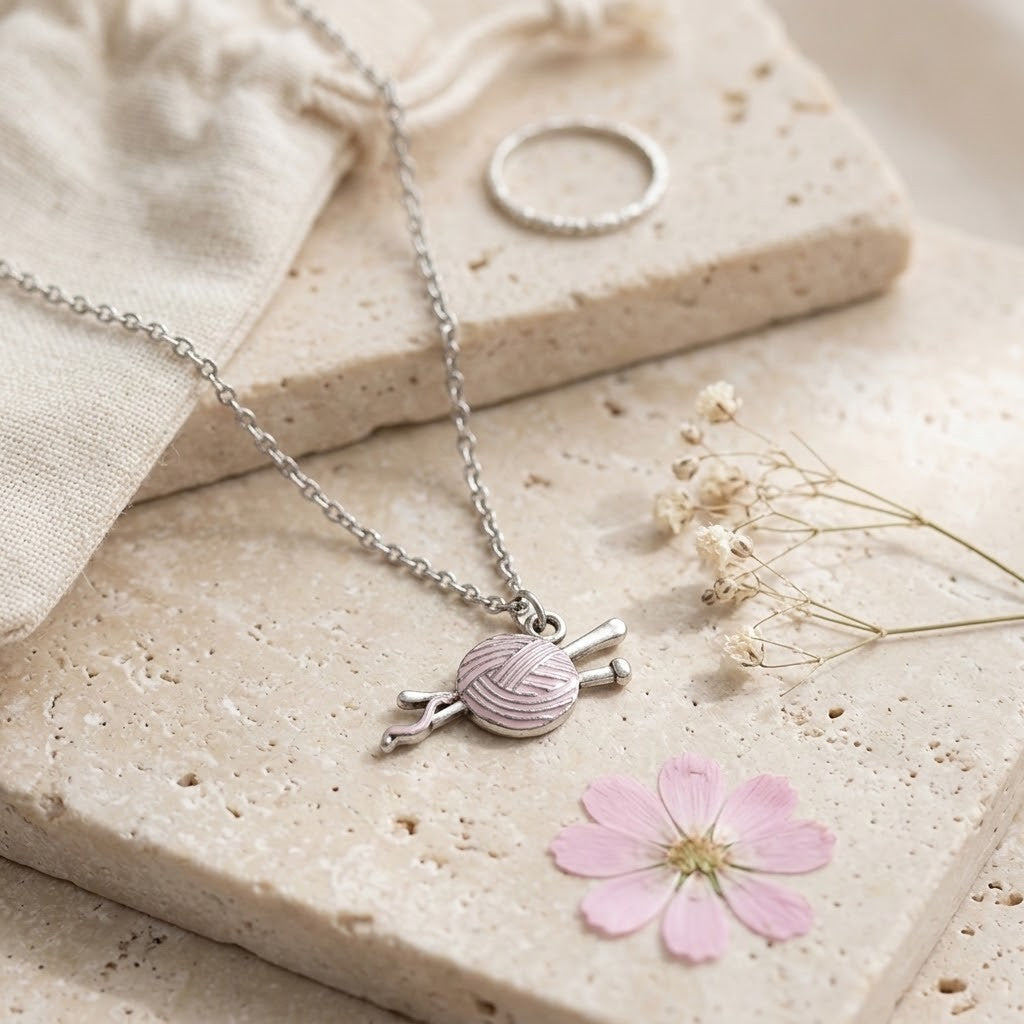 A silver necklace with a knitting needle and yarn charm, styled on beige stone blocks with a small pink flower and a silver ring.