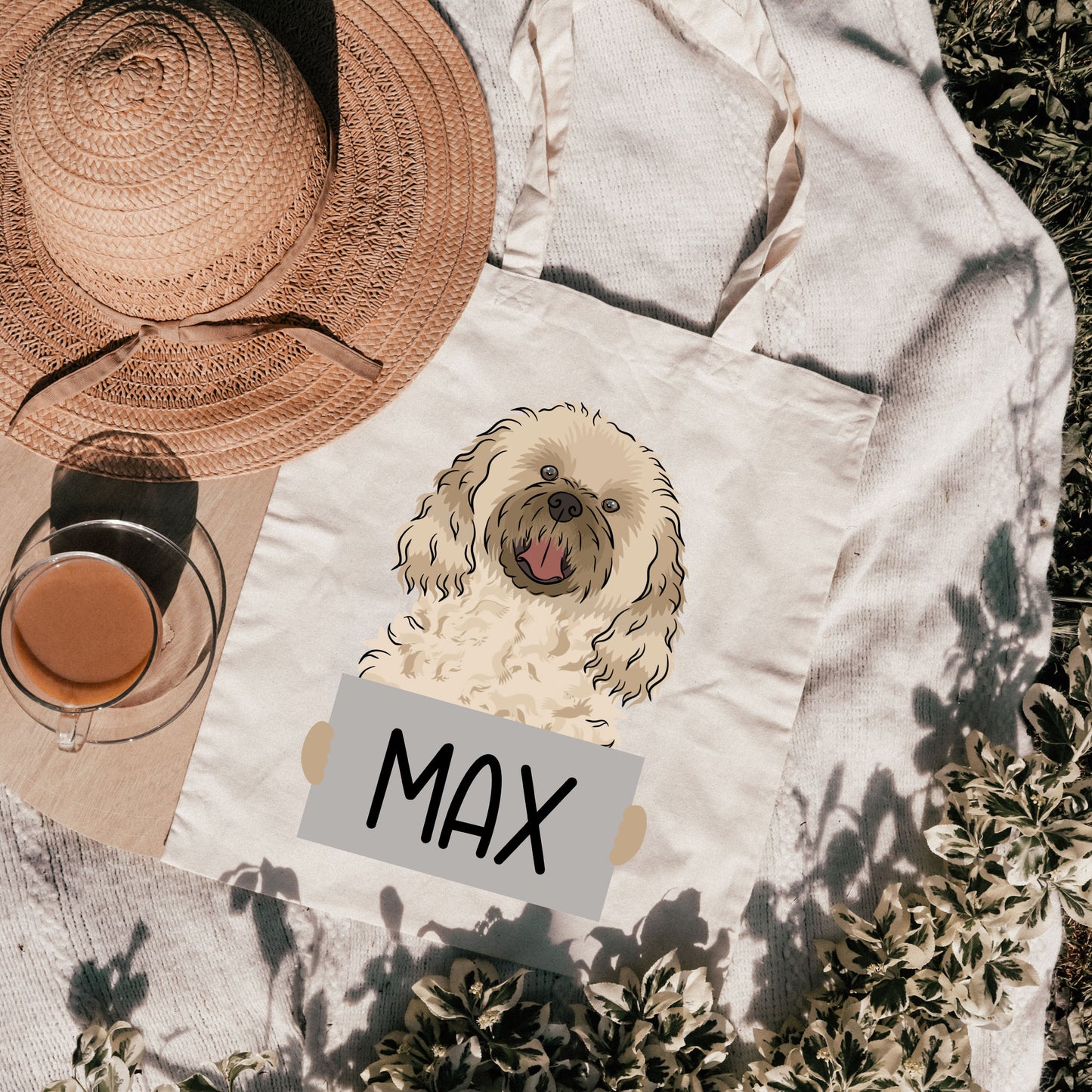 A white cotton tote bag with a personalized print of a cavachon dog named 'Max' on the front, with a green field and a straw hat in the background.