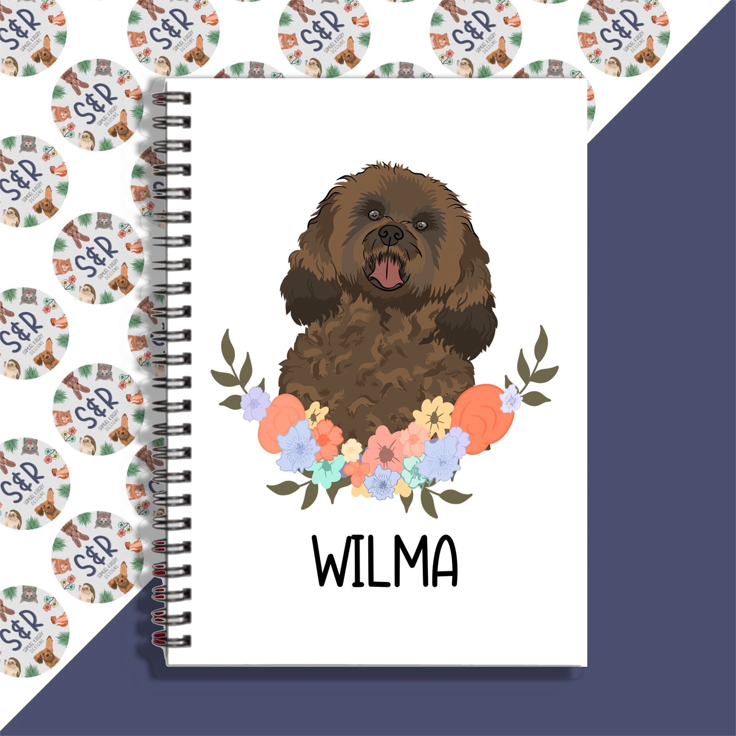 A personalised notebook with an illustration of a cavapoo dog on the cover, surrounded by a floral pattern, with the name 'Wilma' written on the front.