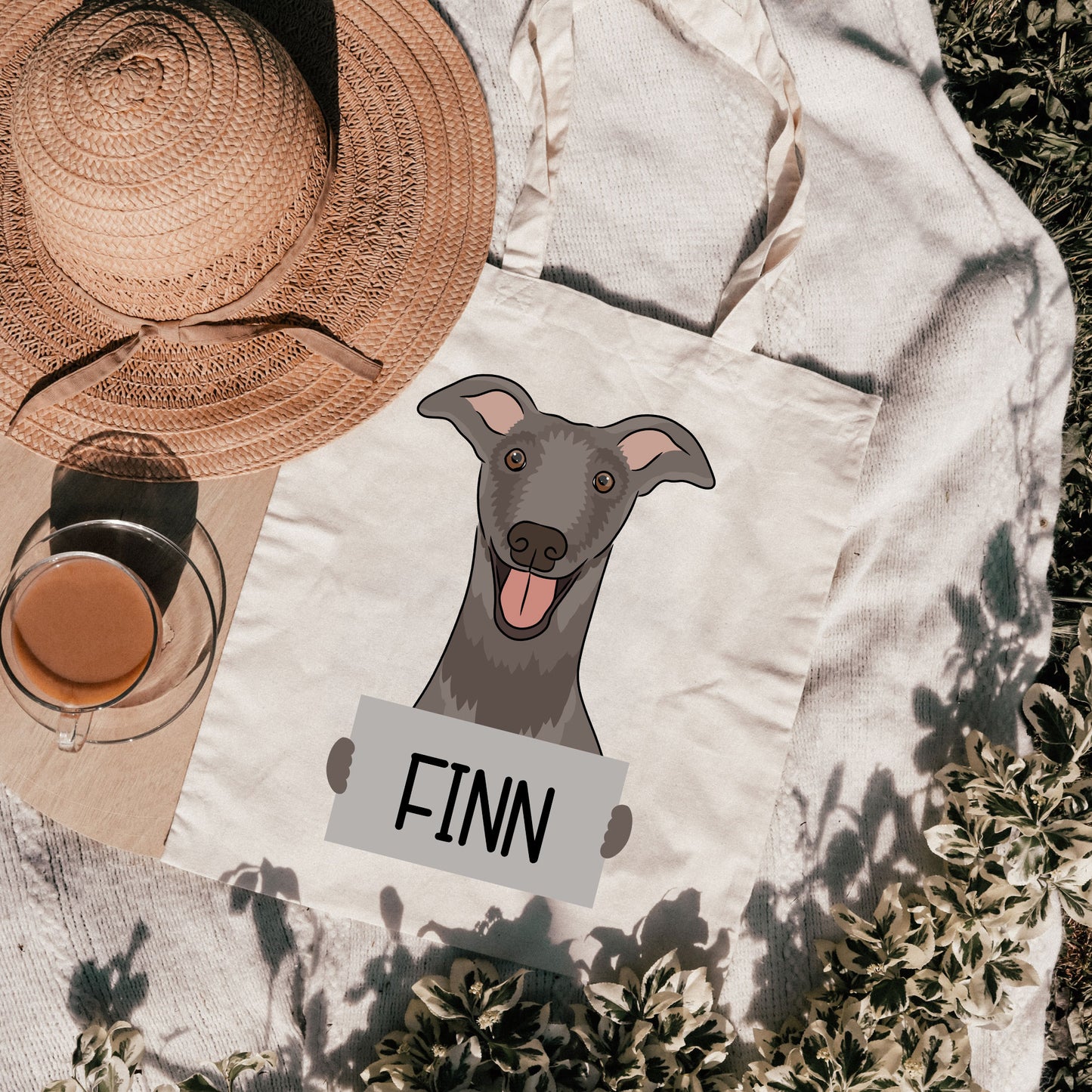 A personalised tote bag featuring a graphic of a greyhound dog with the name 'FINN' printed on it, placed on a grassy surface with a hat and a cup in the background.