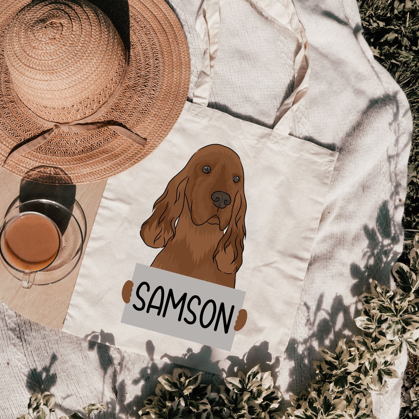 A white polyester tote bag with a personalised graphic print of an Irish Setter dog and the name 'Samson' on it, displayed outdoors with a straw hat and a glass in the background.