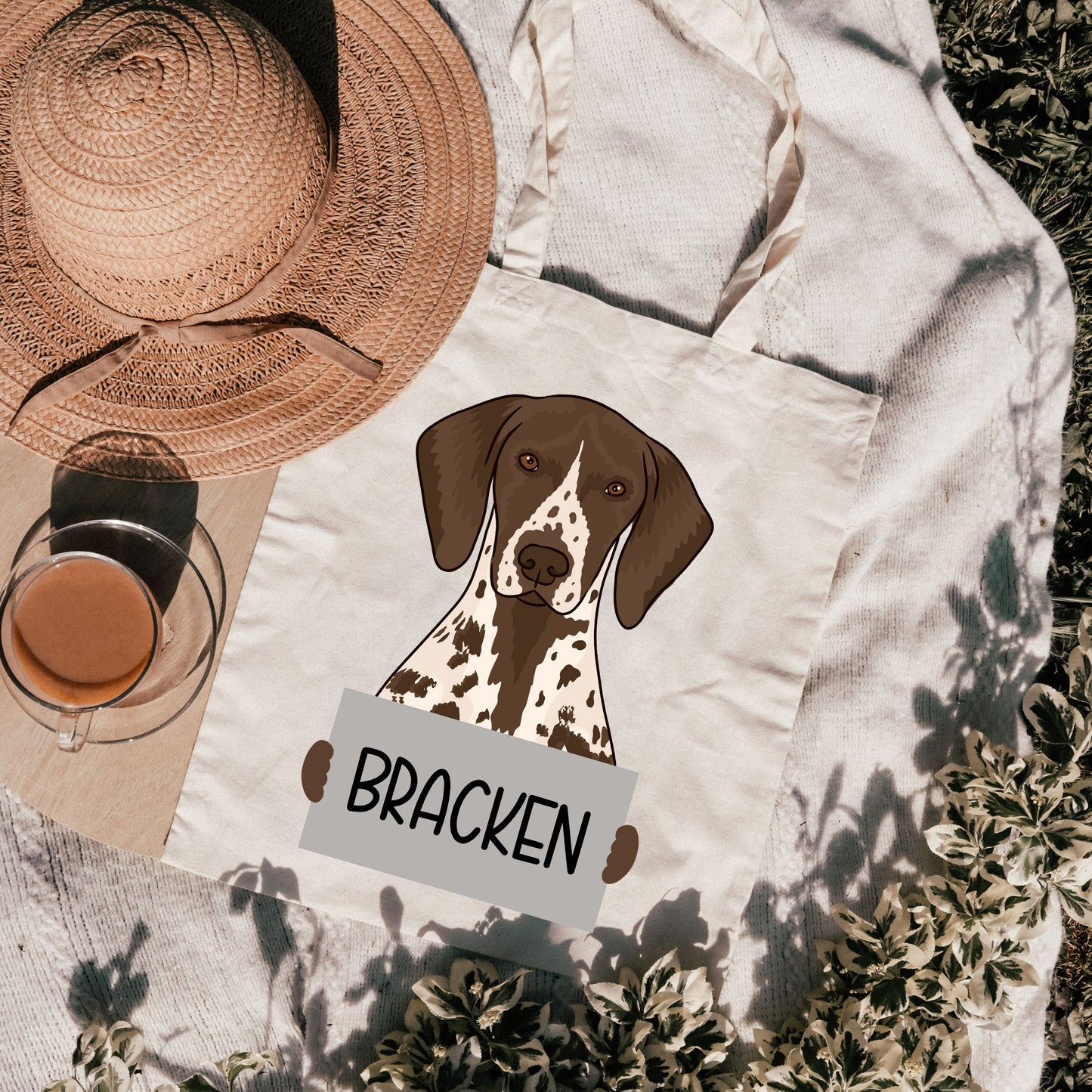 A personalised tote bag with an illustration of a dog breed, named 'Bracken', on a beige background with a hat and a glass of tea in the background.