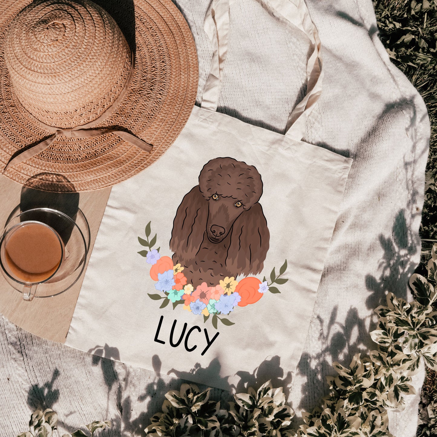 A personalised tote bag featuring an illustration of a poodle dog with a floral design and the name 'LUCY' printed on it, placed on a grassy surface with a hat and a cup in the background.