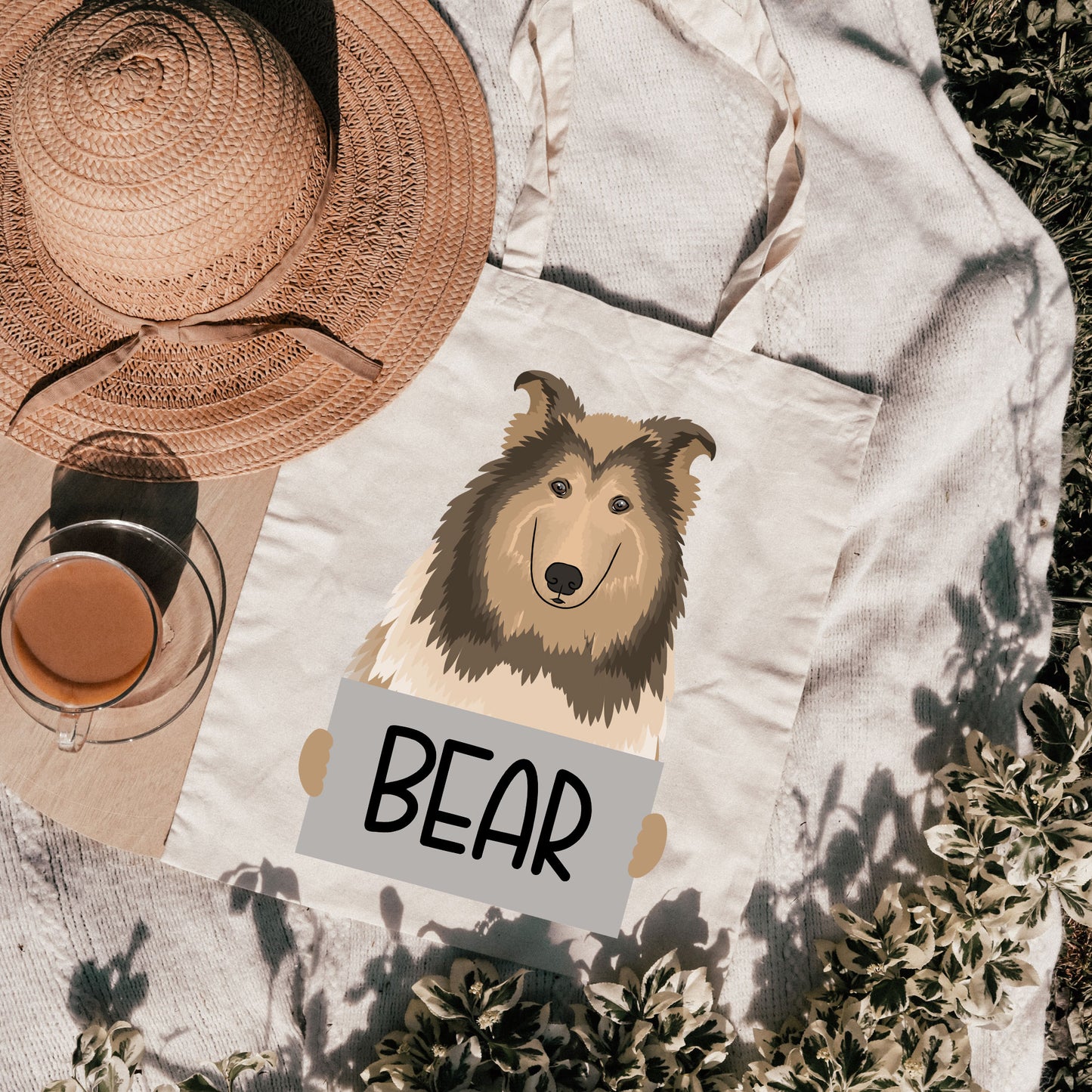 A canvas tote bag with a printed image of a Rough Collie dog breed, with the text 'BEAR' below it, on a outdoor background with a hat and a glass in the background.