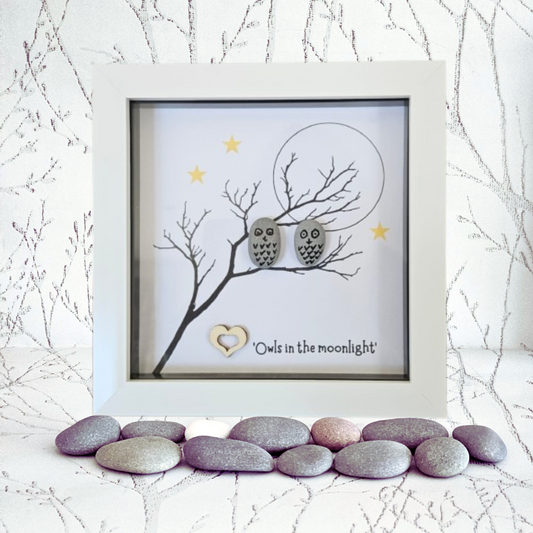 Framed artwork with two owls on a branch, surrounded by pebbles and branches in the foreground.