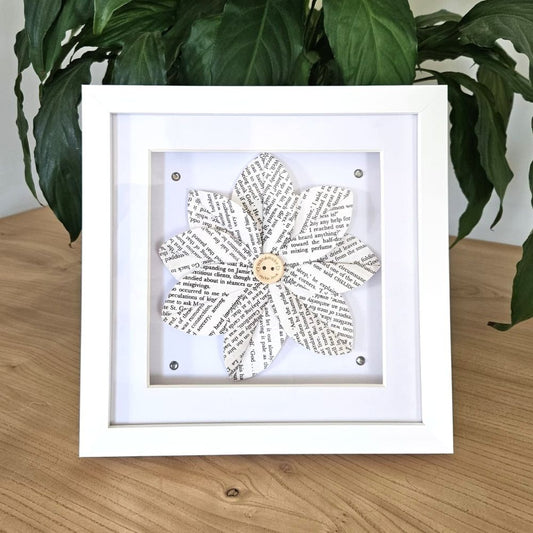 Framed paper flower art on a wooden surface with plants in the background