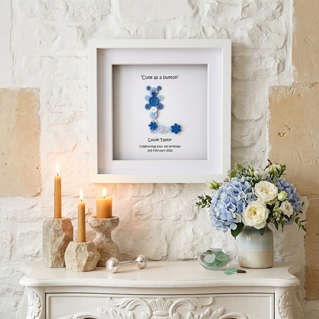 A framed custom keepsake print featuring a blue button letter 'L' with the caption 'Cute as a button' and personalised text for Louie Taylor, displayed on a white brick wall above a console table with candles and flowers.