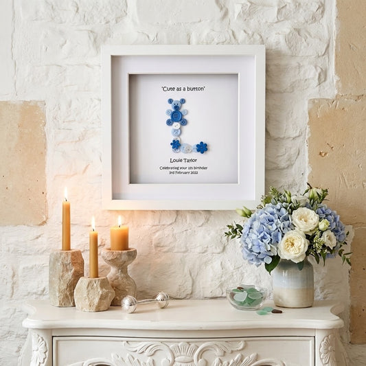 A framed custom keepsake print featuring a blue button letter 'L' with the caption 'Cute as a button' and personalised text for Louie Taylor, displayed on a white brick wall above a console table with candles and flowers.