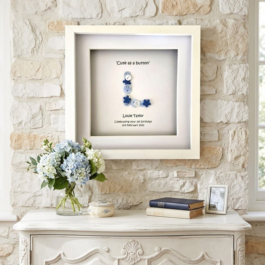 A personalised framed shadow box print hanging on a light stone wall above a white console table. The frame contains a white background with a blue and white decorative letter 'L', the text 'Cute as a button', and the names 'Louise Taito' with dates.